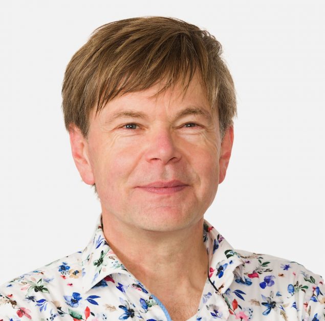a white man with honey coloured hair wears a colourful shirt and smiles at the camera