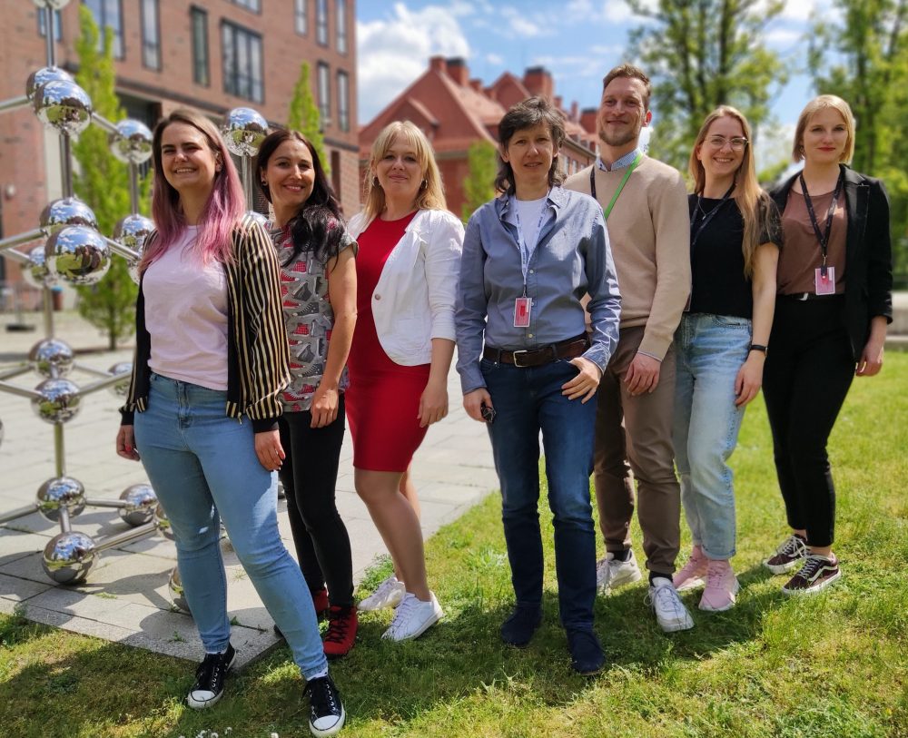 A group of 7 people in casual business clothes stand on a lawn in front of red brick buildings.