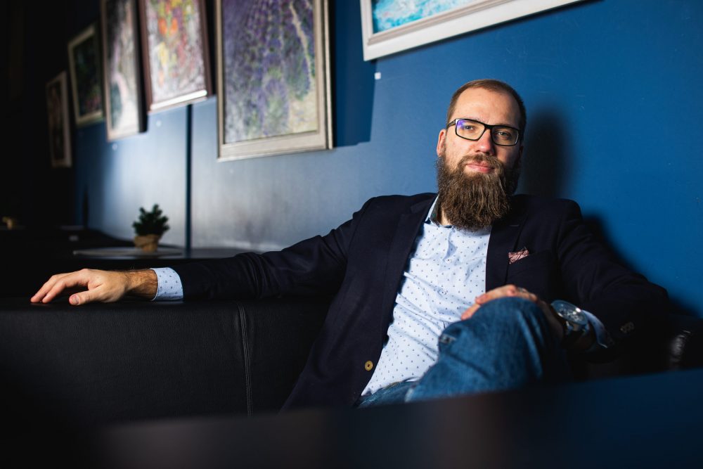 A white man with short brown hair, a beard and glasses is wearing a shirt, jacket and jeans. He is looking at the camera in a relaxed way.