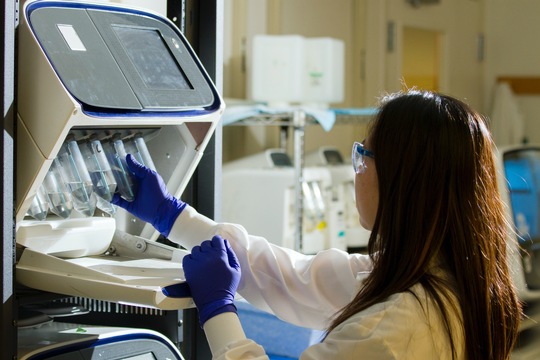 A woman with long dark hair is wearing a lab coat and gloves as she handles samples from a storage facility