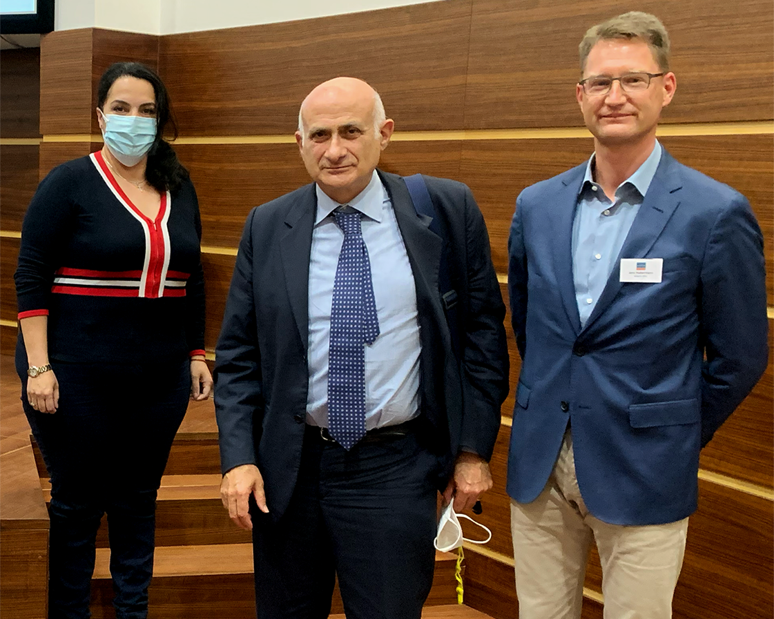 Three people stand together - a woman with long wave dark hair who is wearing black and a face mask, a man wearing a dark suit and carrying a backpack and another man wearing a blue and cream suit and glasses