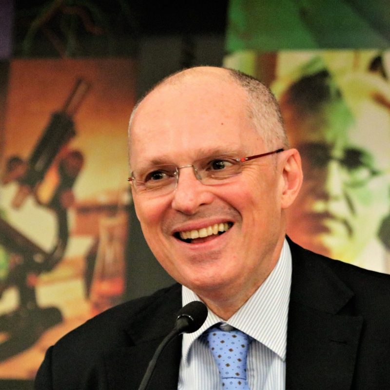 portrait of a white man with short hair and who wears glasses, a suit and tie. He is smiling